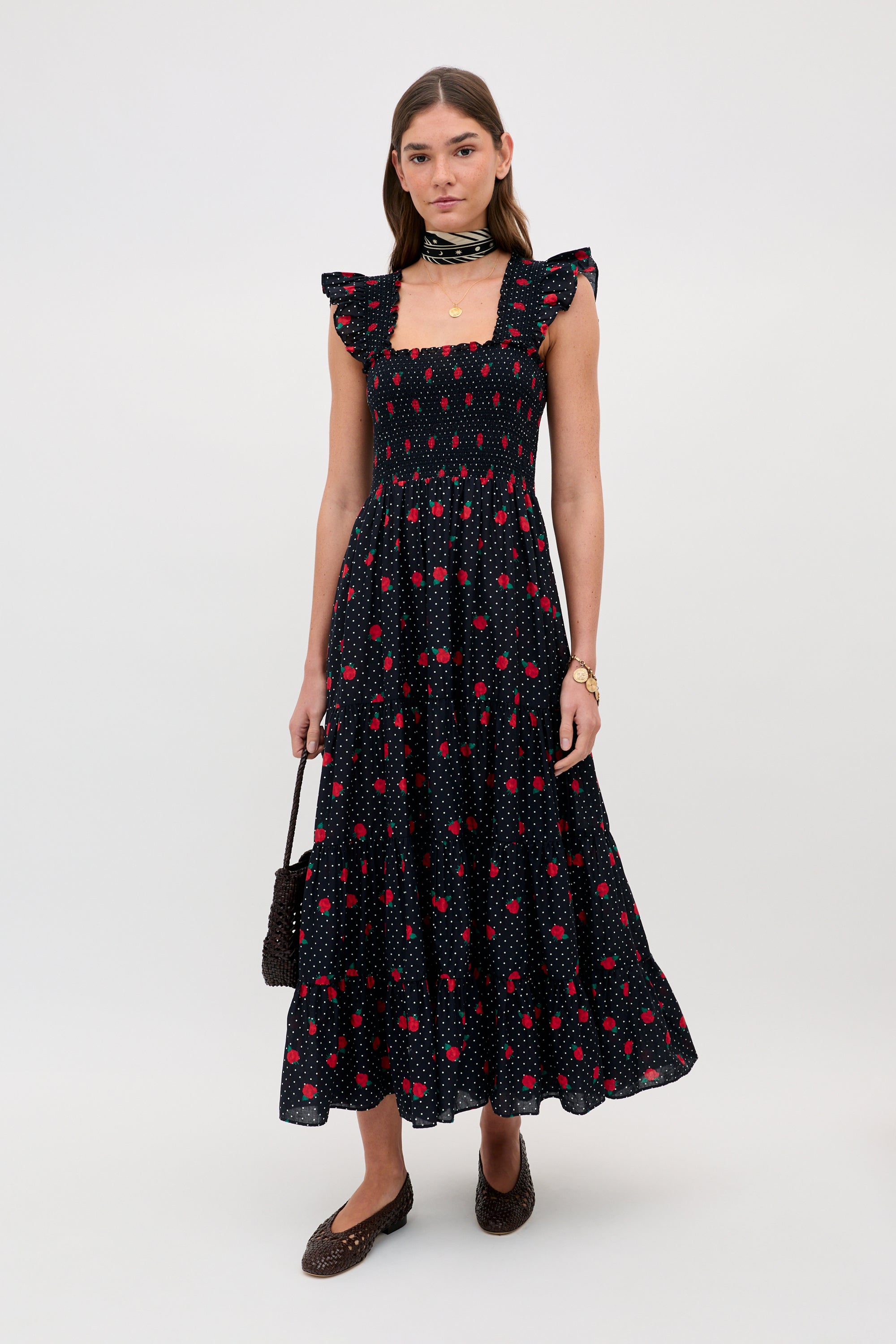 A woman stands against a plain background wearing the Kendall - Rose Spot Black, an ankle-length, sleeveless black dress with red and pink floral patterns, ruffled shoulders, a square neckline, brown flats, a neck scarf, and holding a textured handbag.