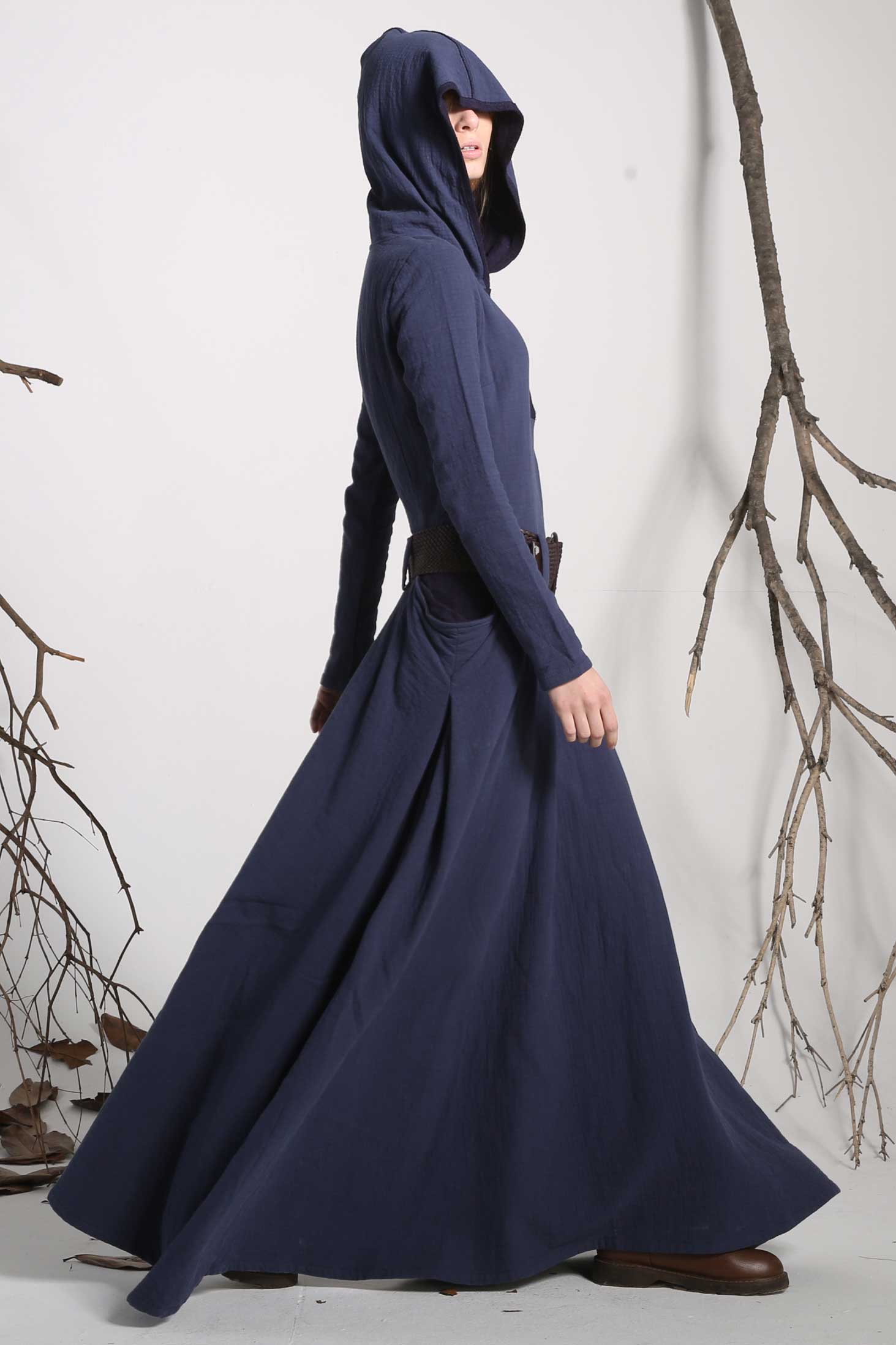 A woman wearing a long, blue hooded linen maxi dress with long sleeves, standing in an outdoor setting with trees in the background.