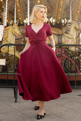 Woman wearing burgundy midi length linen wrap dress with surplice neckline and waist tie standing near carousel