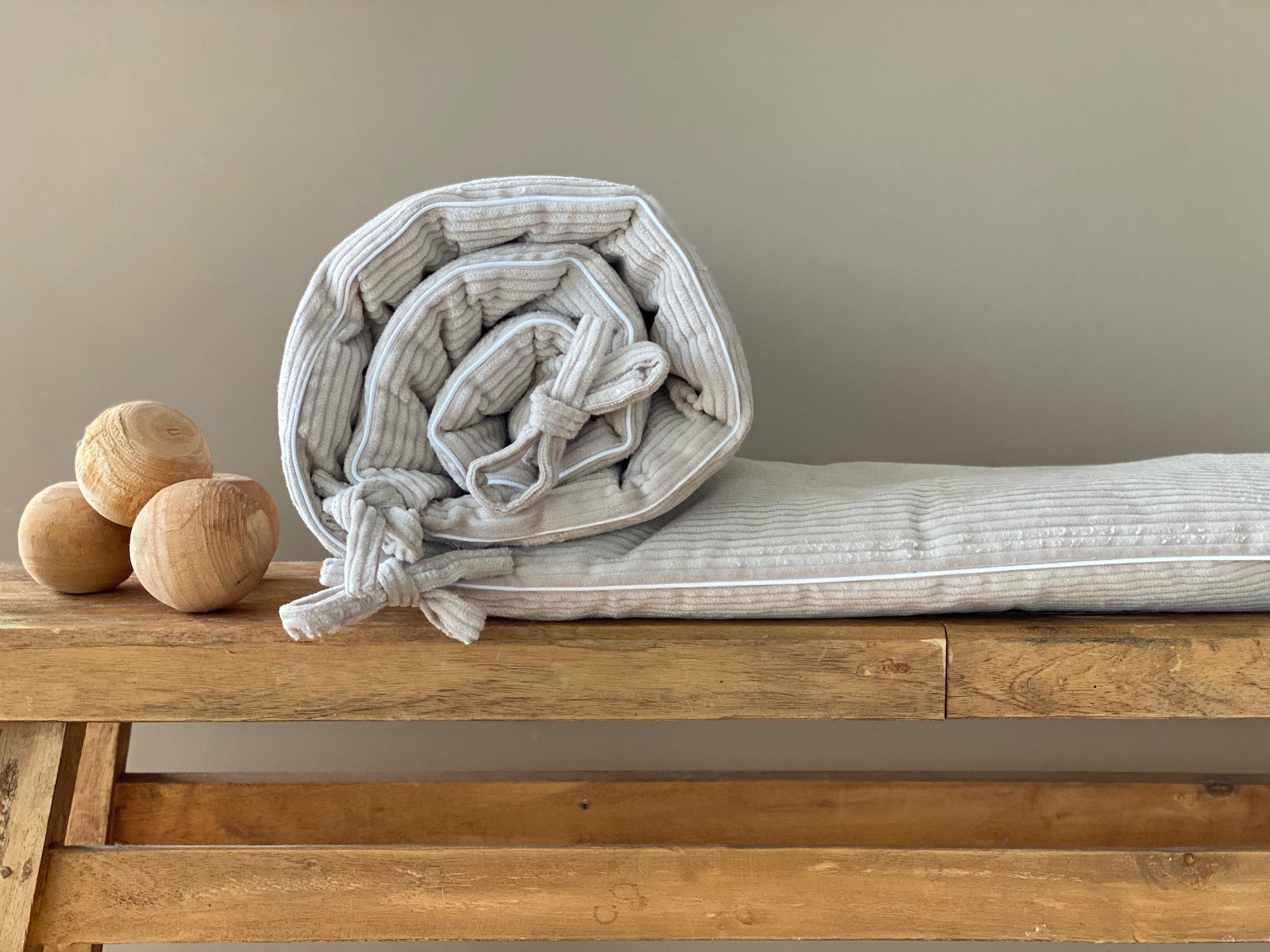 Eine KraftKids Bettumrandung Nestchen Cord Breitcord in Beige Taupe mit Schleifen liegt aufgerollt auf einer Holzbank neben drei glatten, runden Holzkugeln vor einer schlichten beigen Wand.
