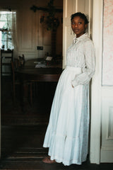 Photo of young woman standing in a doorway wearing prairie dress and apron. model rests back on door frame and has her hands in the pockets of the apron.