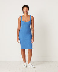 A person stands facing forward in a bright, minimal room, wearing a blue Hunza G Tank Dress—an original crinkle seersucker, one-size, sleeveless and form-fitting—and white sneakers. The space has plain white walls and a white wooden floor.