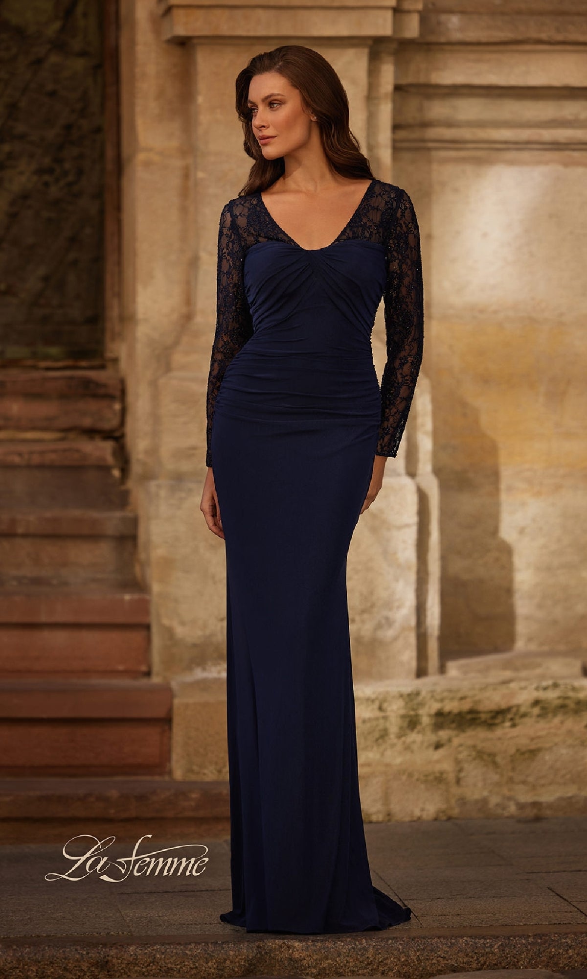 A woman wearing the Long Lace-Sleeve Jersey Formal Dress: La Femme 33121, a fitted navy evening gown with lace sleeves, stands near stone steps and a textured wall.