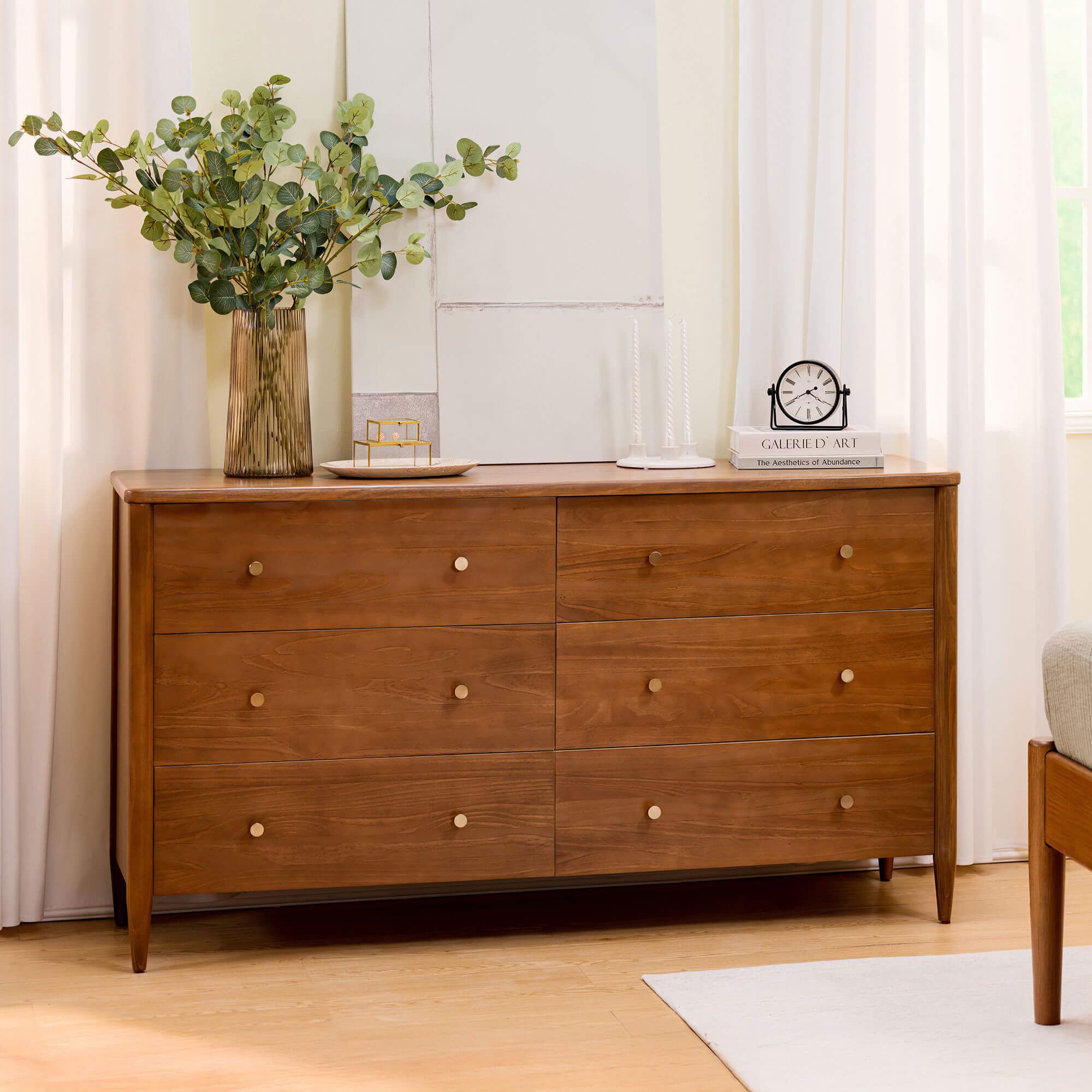 Mid-century modern walnut six-drawer wooden dresser with brass knobs in a bright bedroom, styled with eucalyptus vase, clock and books