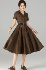 A woman wearing a brown button-up linen knee-length dress with short sleeves, paired with a belt and standing against a white background.