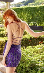 A woman with long red hair wears the Starburst Beaded Backless Short Party Dress 4001, a strappy purple sequin cocktail dress, in a garden setting. "PRIMAVERA" appears on the image.