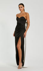 A woman in a Mac Duggal 49879 Strapless Long Bow Prom Dress with a thigh-high slit, silver heels, and her hair pulled back stands against a plain light background.