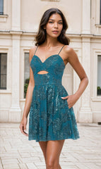 A woman stands outdoors wearing the Cut-Out Short Teal Glitter-Print Hoco Dress 4997BN, featuring spaghetti straps, a waist cutout, and sparkling embellishments. She has long wavy hair and a neutral expression.