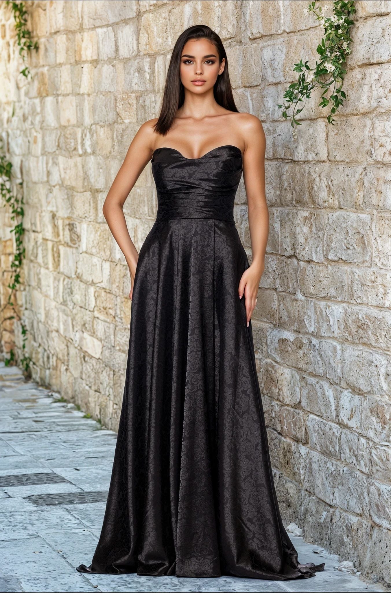 A woman stands against a stone wall, wearing the Strapless Black Prom Gown: Blondie Nites 5301BN—a long black dress with a subtle textured pattern, available in junior sizes for the perfect fit.