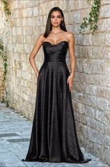 A woman stands against a stone wall, wearing the Strapless Black Prom Gown: Blondie Nites 5301BN—a long black dress with a subtle textured pattern, available in junior sizes for the perfect fit.