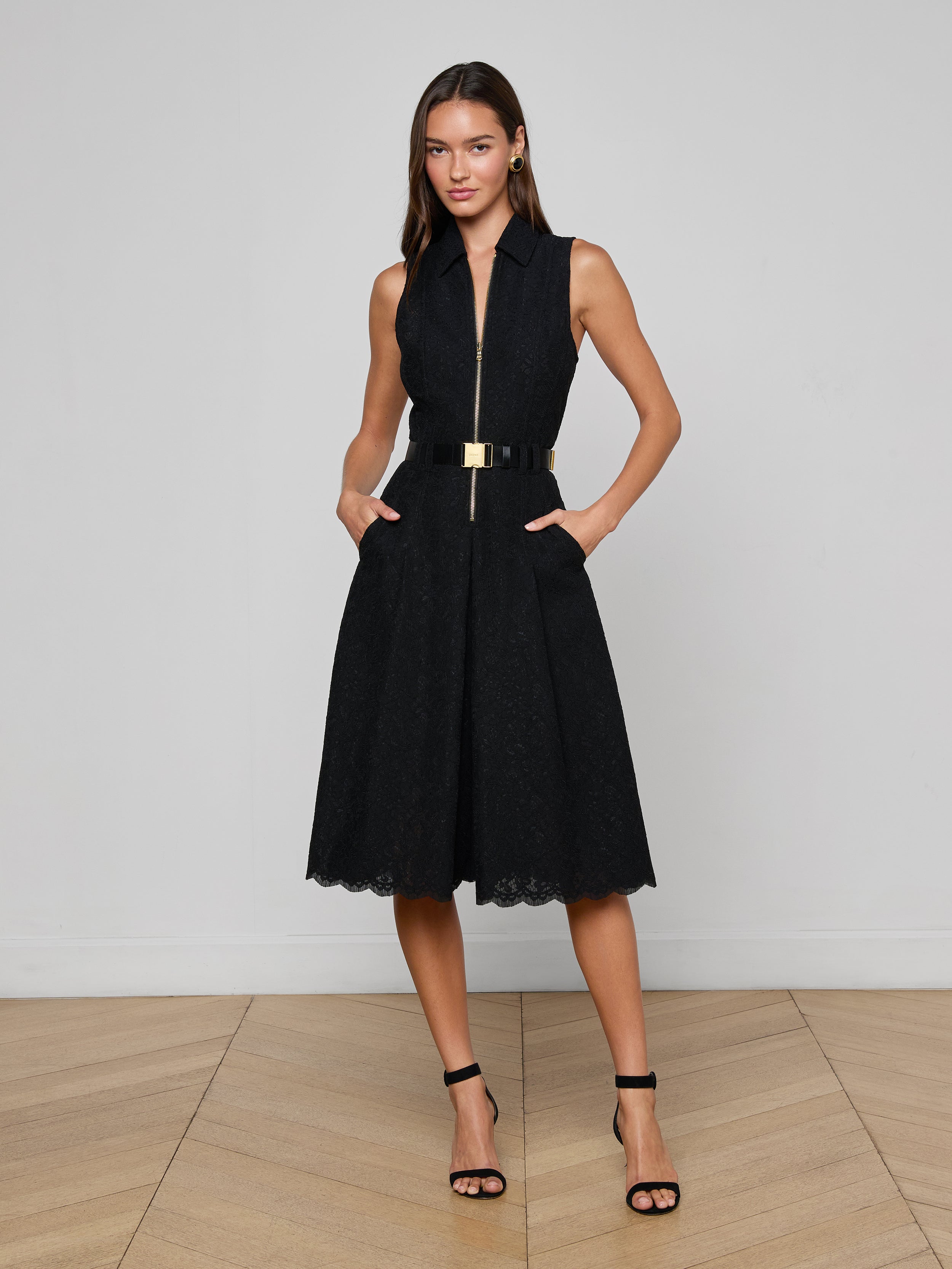 A woman stands on a wooden floor against a white wall, wearing the Kylo Lace Shirt Dress with a zip front, black belt with gold buckle, black strappy heels, and her hands casually in the dress’s pockets.