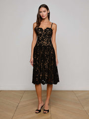 A woman with long, wavy brown hair poses indoors against a light wall and wooden floor, wearing the L'AGENCE Sale Flyn Lace Midi Dress— a sleeveless black floral eyelet lace dress with a scalloped neckline—paired with black heels.