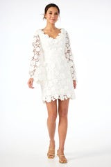 A woman wearing the Ariah Long-Sleeve Dress - Love All Lace in Whitewash, featuring sheer floral sleeves and scalloped edges, paired with gold strappy heels, stands against a plain white background.