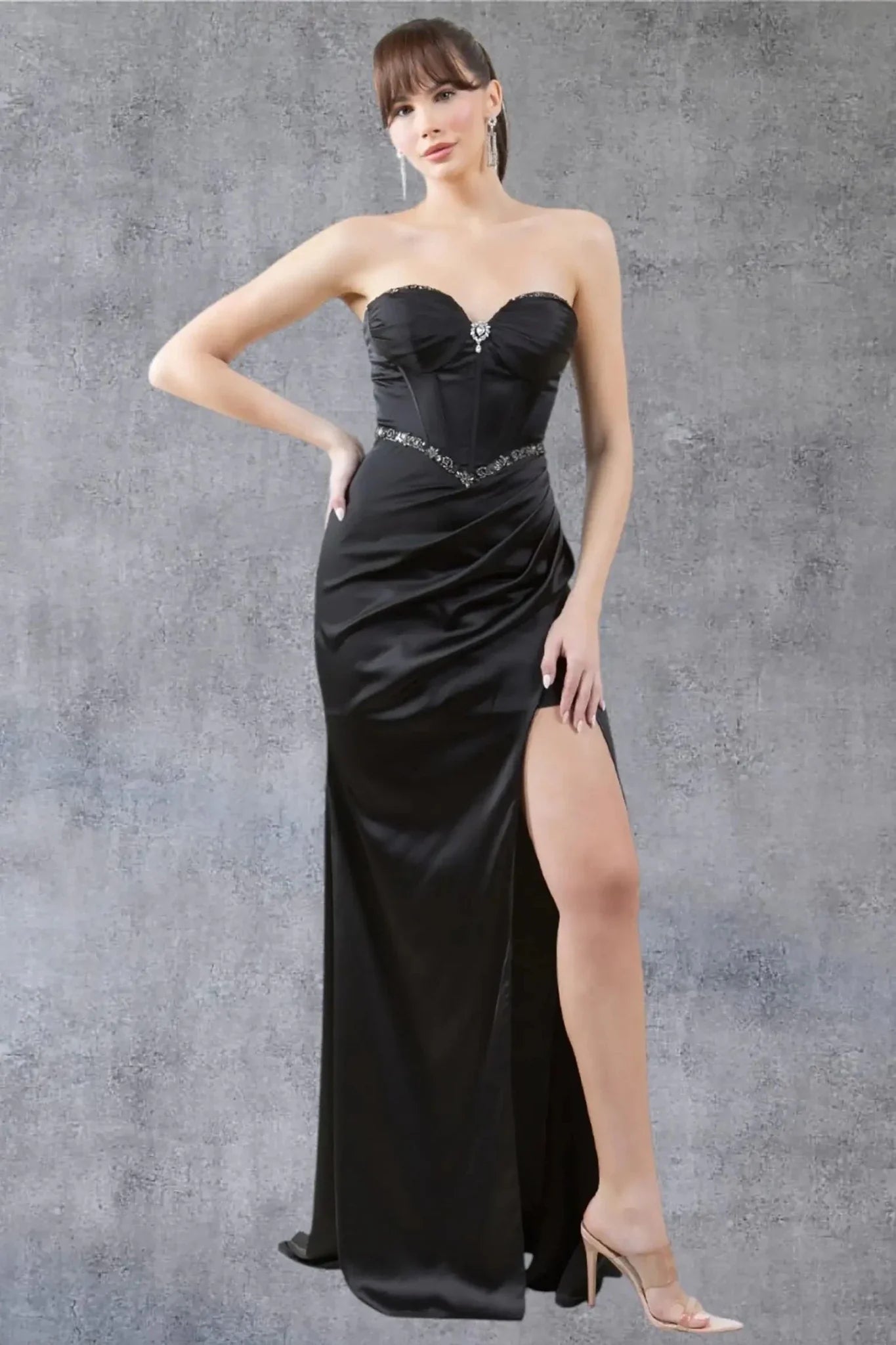 Full front view of a women wearing black evening prom dress with strapless corset bodice, brooch detail, and thigh-high slit.