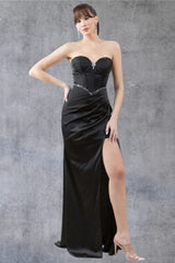 Full front view of a women wearing black evening prom dress with strapless corset bodice, brooch detail, and thigh-high slit.