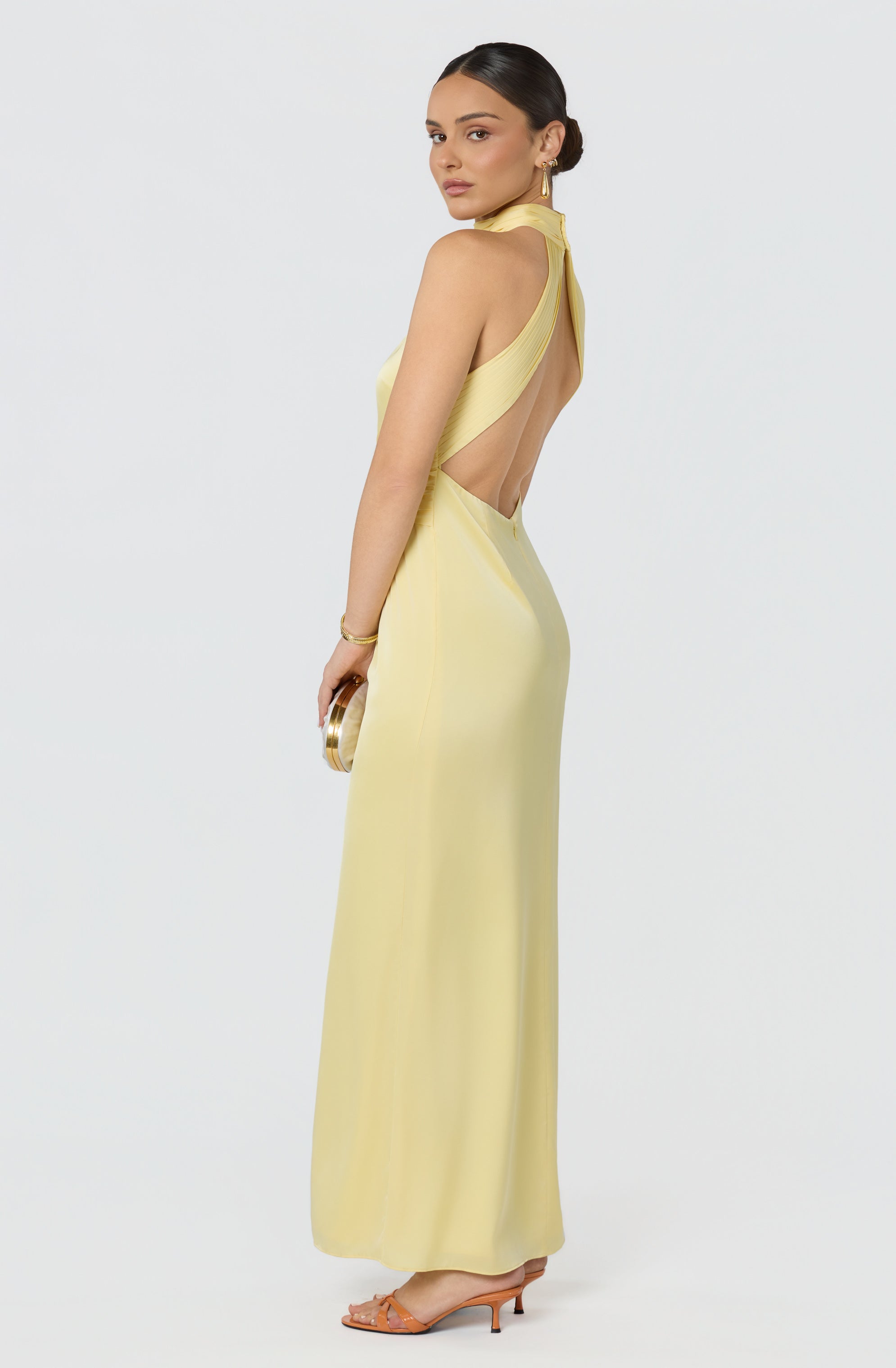 Woman in a long, yellow dress with open back and cutout details, wearing earrings and holding a clutch.