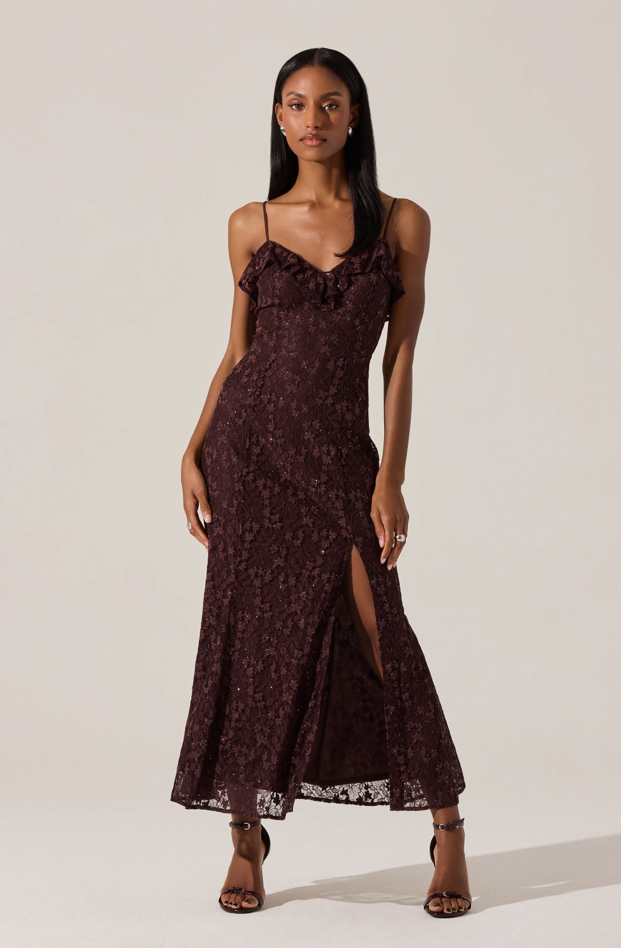 A woman is wearing a long, burgundy lace dress with spaghetti straps and a ruffled neckline. The dress features a high slit on one side, revealing her leg. She is also wearing black open-toe high-heeled sandals.