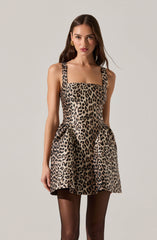 A woman is wearing a sleeveless, knee-length dress with a square neckline and wide shoulder straps. The dress features a bold leopard print pattern in shades of tan, black, and beige. She is also wearing sheer black tights.