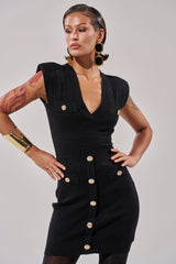 A woman wears the SUSIE SLEEVELESS MINI DRESS IN BLACK, a contoured fit stretch-knit mini dress with gold buttons. She pairs it with large gold earrings, a wide cuff bracelet, and displays tattoos on her left arm. Her hair is short and slicked back.