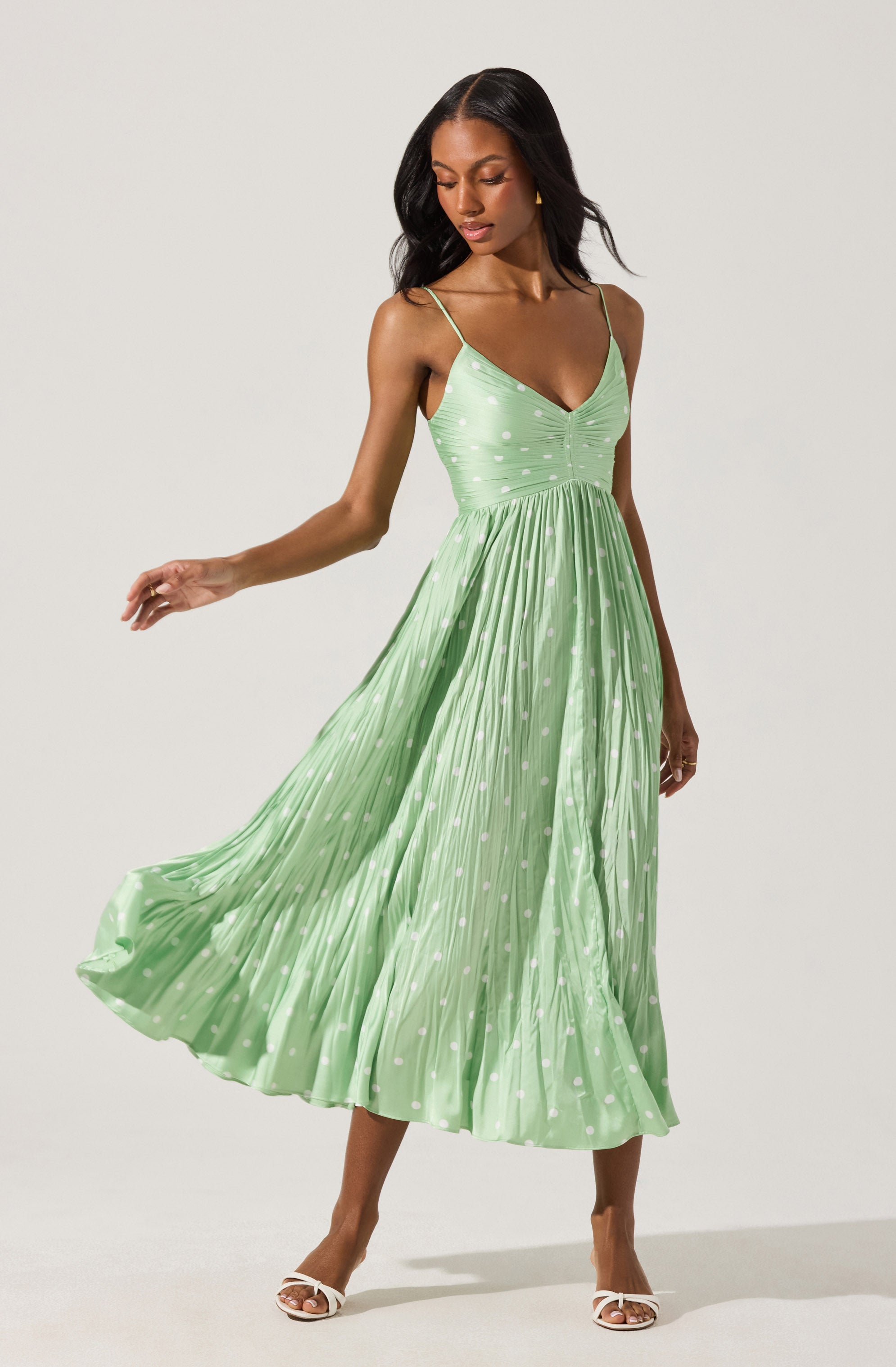 A woman is wearing a light green midi dress with thin spaghetti straps and a V-neckline. The dress features a fitted bodice with a ruched detail at the center and a full, flowing skirt with a pleated texture. The fabric has a subtle polka dot pattern. She is also wearing white open-toe heels.