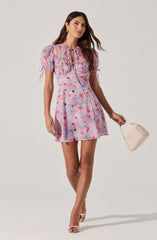 A woman is wearing a colorful, watercolor-like patterned dress with shades of pink, purple, and blue. The dress has puffed short sleeves with adjustable drawstrings, a fitted waist, and a short, above-the-knee hemline. She is accessorizing with gold earrings and rings, and holding a small beige handbag. She is also wearing light pink open-toe heels.
