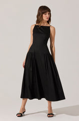 A woman is wearing a sleeveless, ankle-length black dress with a fitted bodice and a full, gathered skirt. She has on black high-heeled sandals with decorative bows on the front.