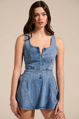 A woman with long brown hair and large hoop earrings stands against a beige background in the VIVIAN DENIM MINI DRESS, a sleeveless mini dress featuring a zipper front, corset bodice for structure, and stretchy fabric for comfort.