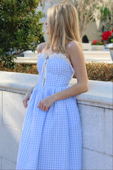 A woman wearing a blue and white gingham maxi dress with lace trim on the bodice. The dress features a corset-style tie at the front and appears to be sleeveless.
