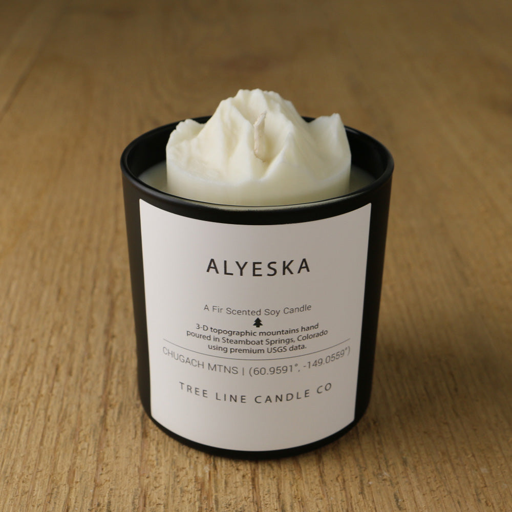 A white soy wax replica candle of Alyeska in a round, black glass.