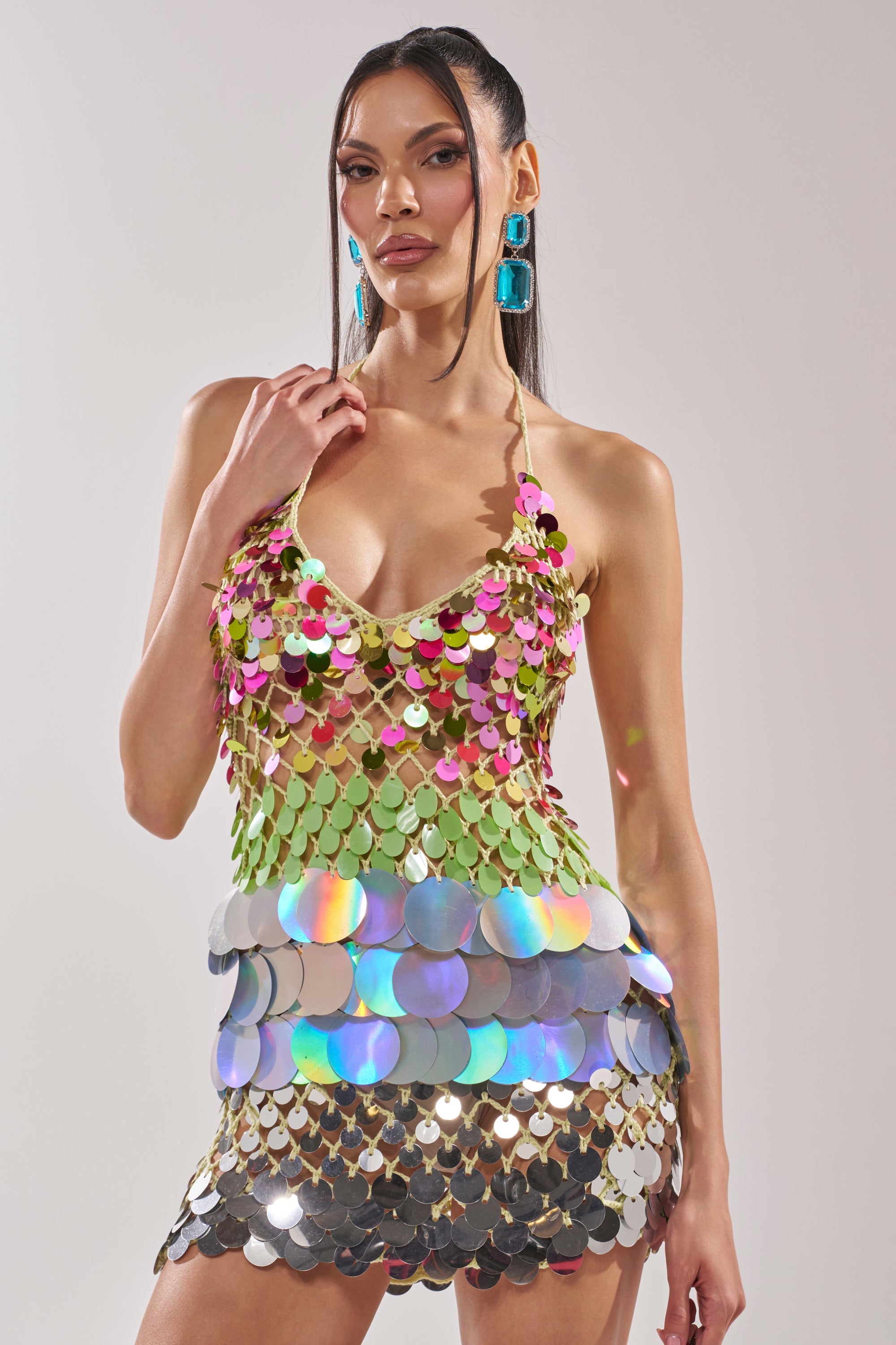 A woman poses in the MAKE IT TO THE MORNING CROCHET MINI DRESS, a halter mini dress with large multicolored sequins, deep neckline, and iridescent discs at the hips. She styles it with striking blue rectangular earrings and sleek, straight hair.