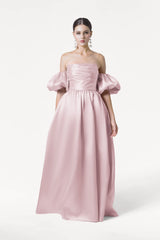 A woman wears the TH&TH Blair Bridesmaid Dress in Blush Pink Satin Twill, featuring voluminous detachable puff sleeves, a fitted bodice, and a long flowing skirt. Her hair is pulled back and she accessorizes with large dangling earrings.