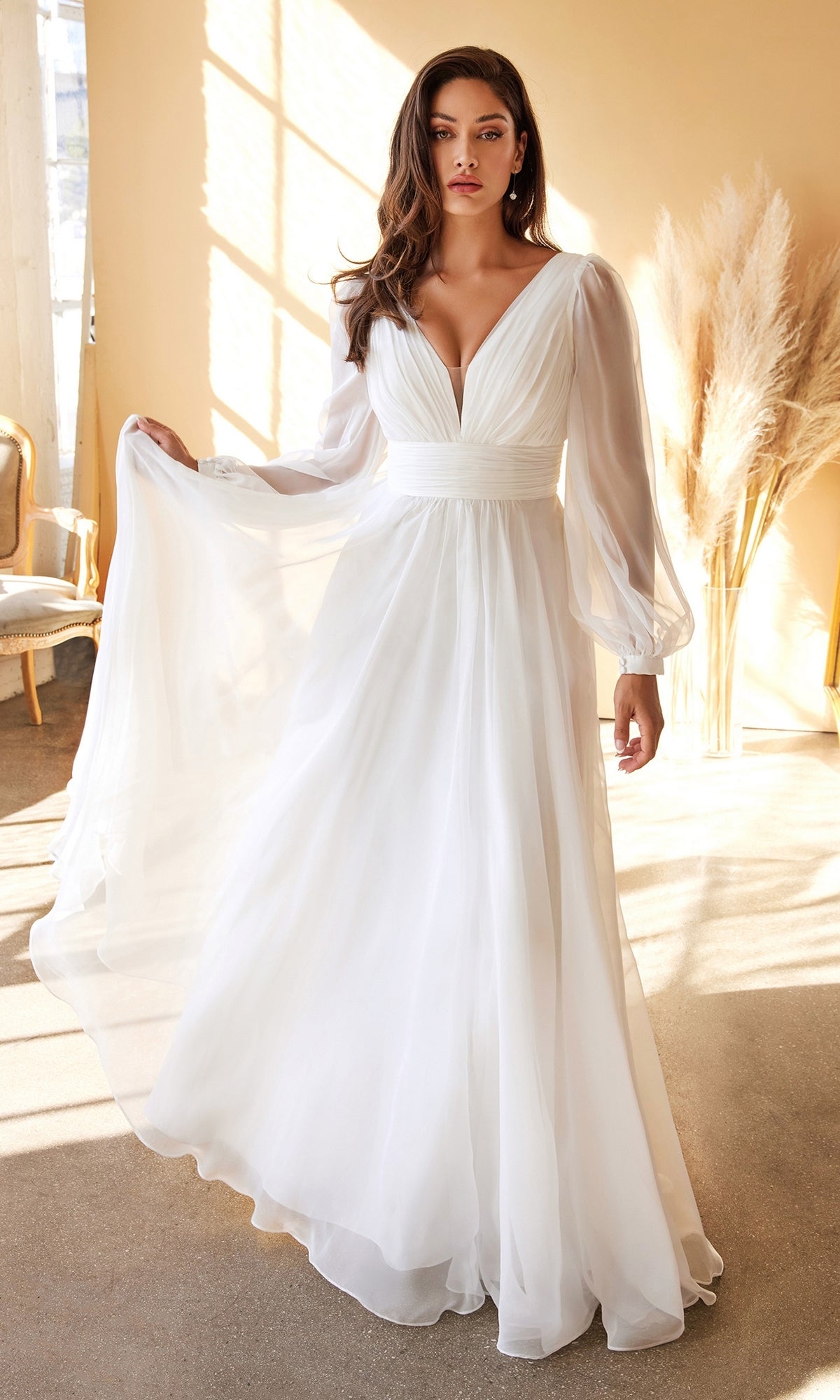 A woman stands indoors near a window and pampas grass, gracefully holding out the skirt of a Long White Chiffon Formal Dress with Long Sleeves.