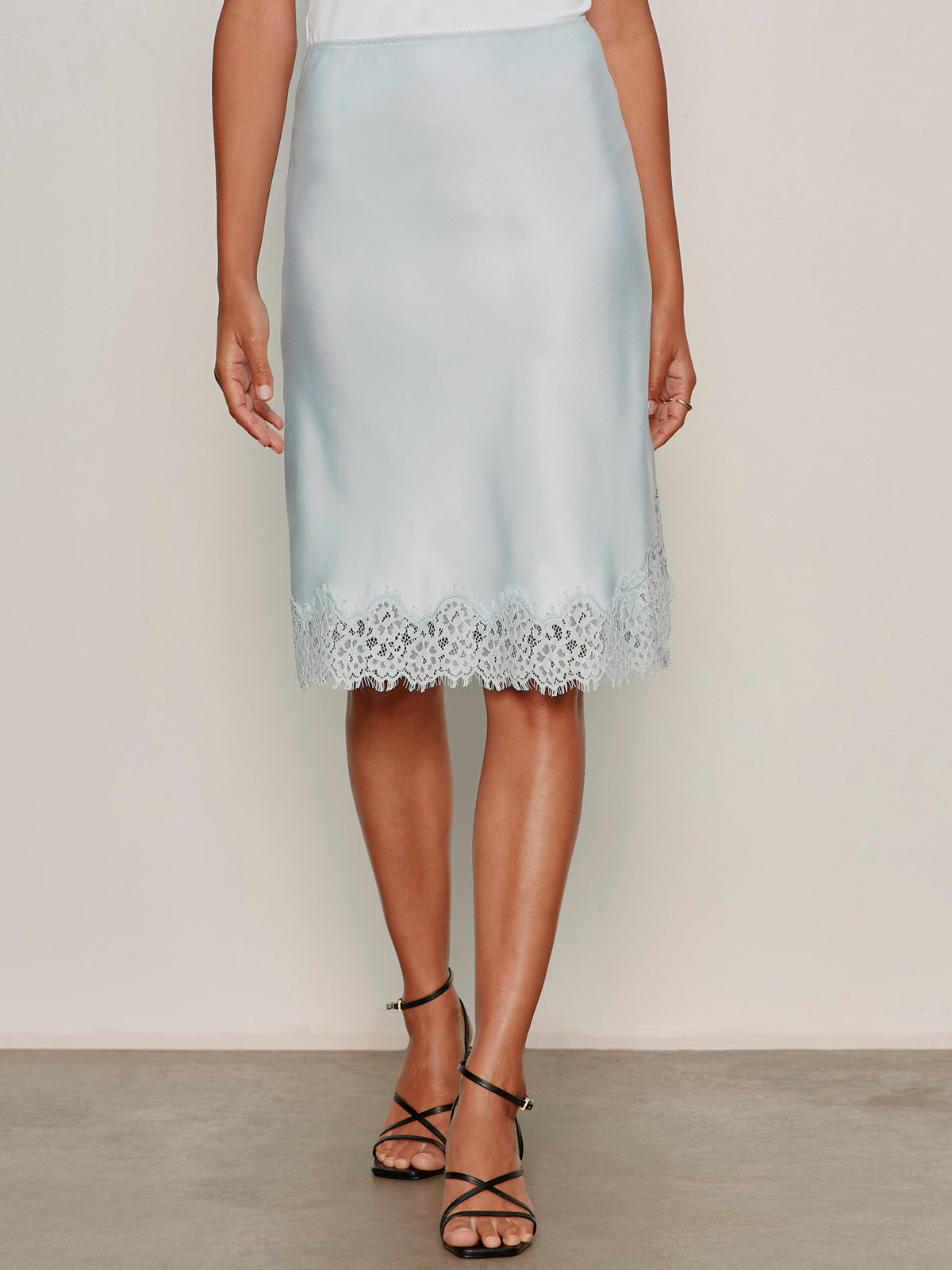 A person wears the Sanctuary Clothing effortless lace slip skirt in blue aura with black strappy heels, standing against a neutral background.