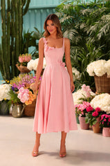 A woman in a soft blush Camellia A-line midi dress stands indoors holding a bouquet, surrounded by potted plants and blooming flowers.