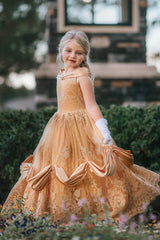 A young girl with blonde hair stands outdoors, wearing the Portrait Collection Red Rose Princess Gown, an intricately designed gold princess dress adorned with floral lace patterns. The satin off shoulder neckline is adorned with a gold jewel.