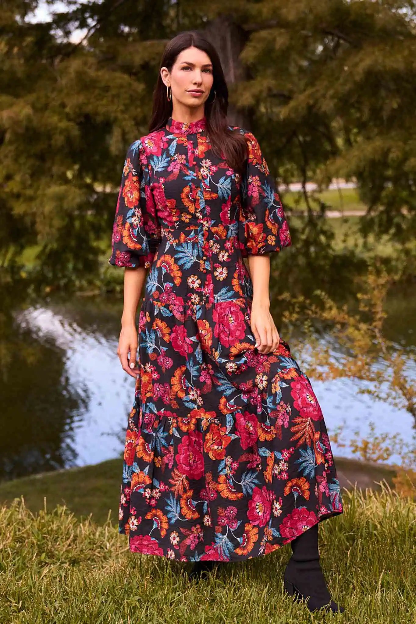 Woman in a park wearing the Desi Floral Maxi Dress with bold floral print, high neck, puff sleeves, styled with hoop earrings and black ankle boots.