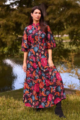Woman in a park wearing the Desi Floral Maxi Dress with bold floral print, high neck, puff sleeves, styled with hoop earrings and black ankle boots.