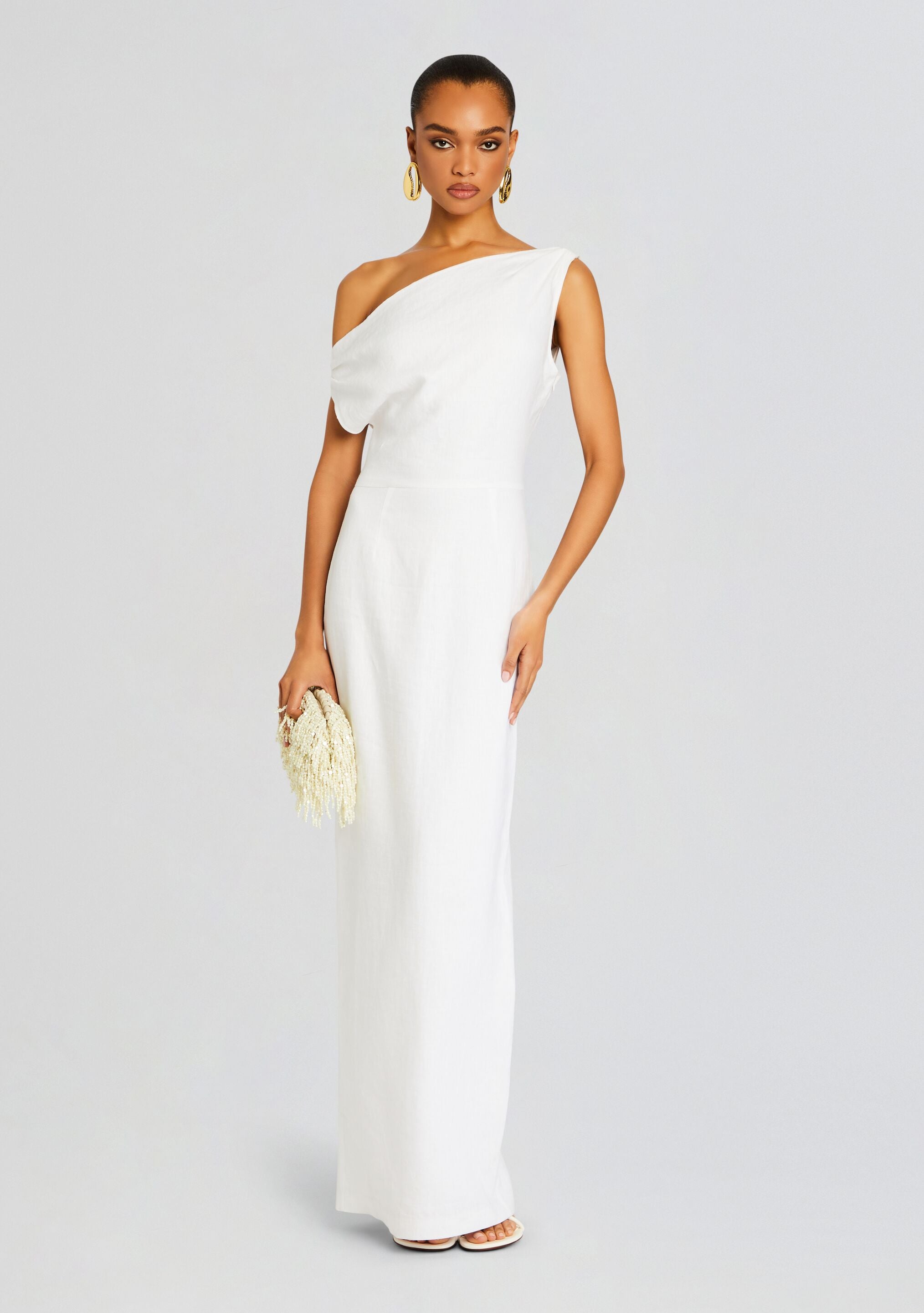 Full-length white linen off-shoulder maxi dress on a model, holding a cream beaded clutch.