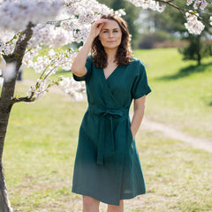 A woman with brown hair, wearing a dark green, short-sleeved wrap dress, stands outdoors under a tree with white blossoms.
