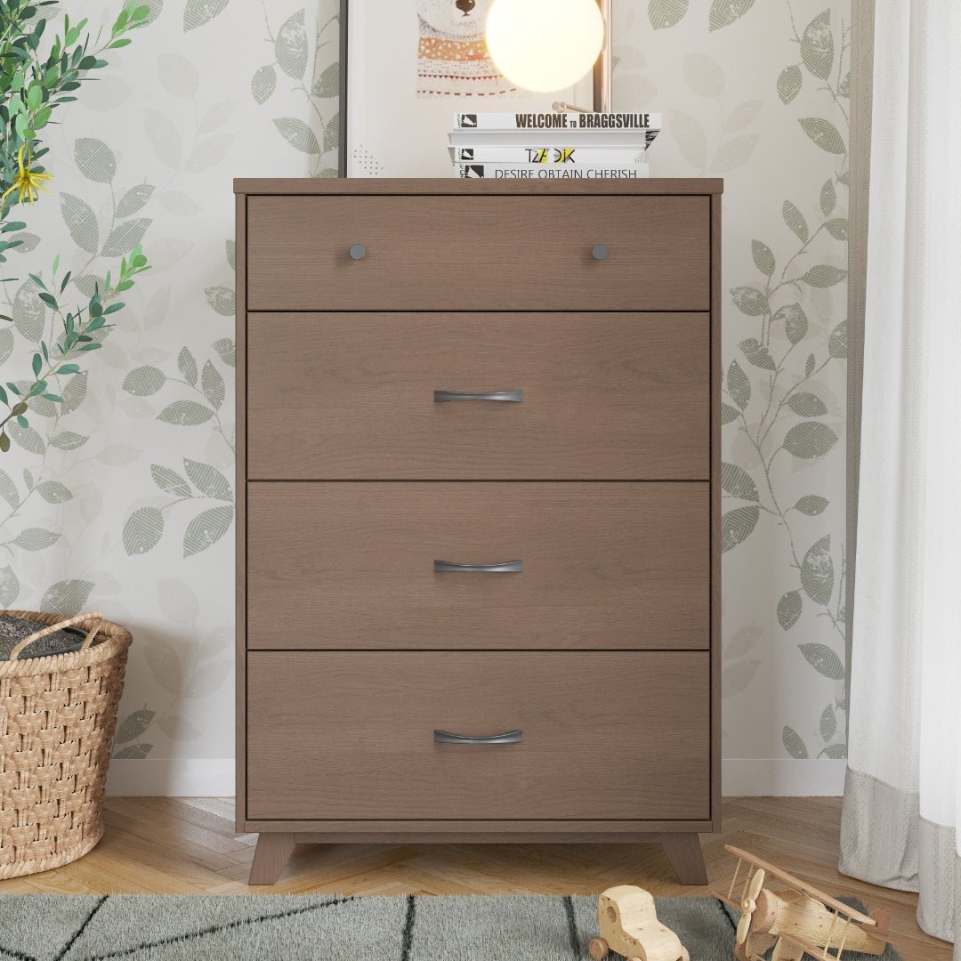 The Child Craft Soho 4 Drawer Chest in Dusty Heather stands against a leafy wallpapered wall, topped with decor, books, and a round lamp. A woven basket and toy wooden train add charm to this stylish mid-century modern nursery setup.