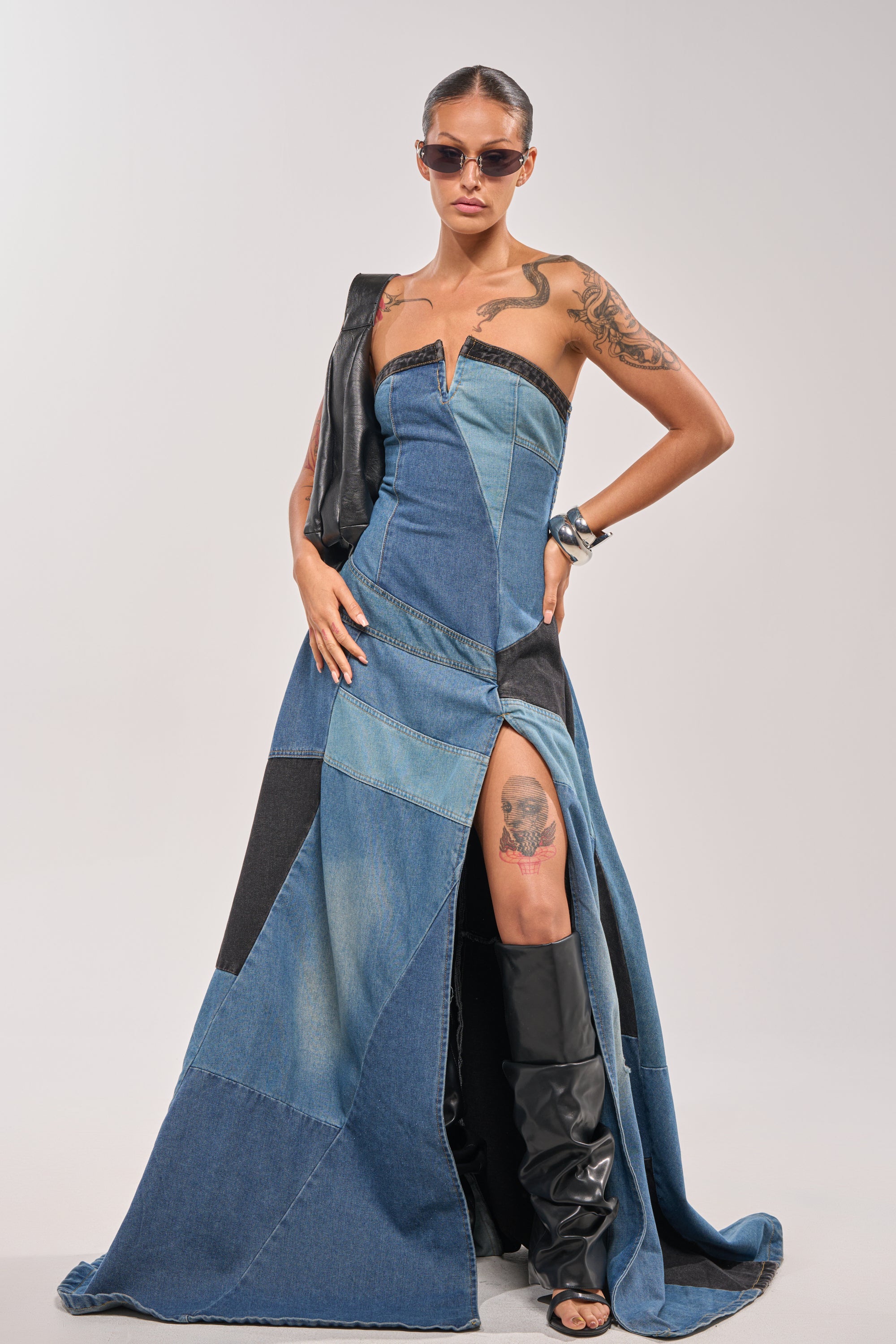 A person poses confidently in the LIFE IN DENIM STRAPLESS MAXI DRESS, featuring patchwork details and a high slit. Styled with black heeled boots, sunglasses, and sleek hair, this look is completed by a black leather accessory over one shoulder.