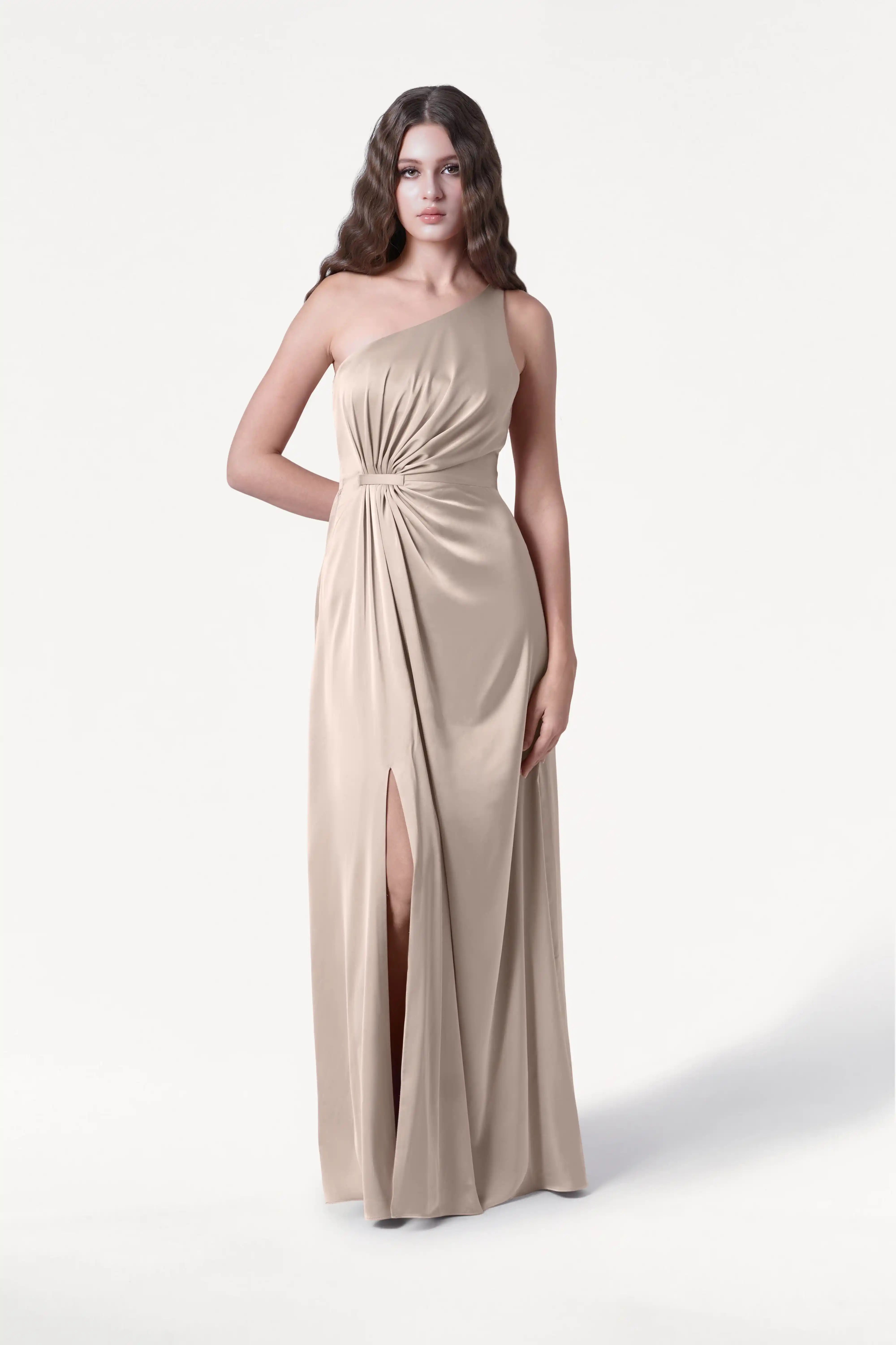 A woman wears the TH&TH Greta Bridesmaid Dress in Champagne Sky Satin, a one-shoulder gown with a gathered waist and thigh-high slit, standing against a plain white background.