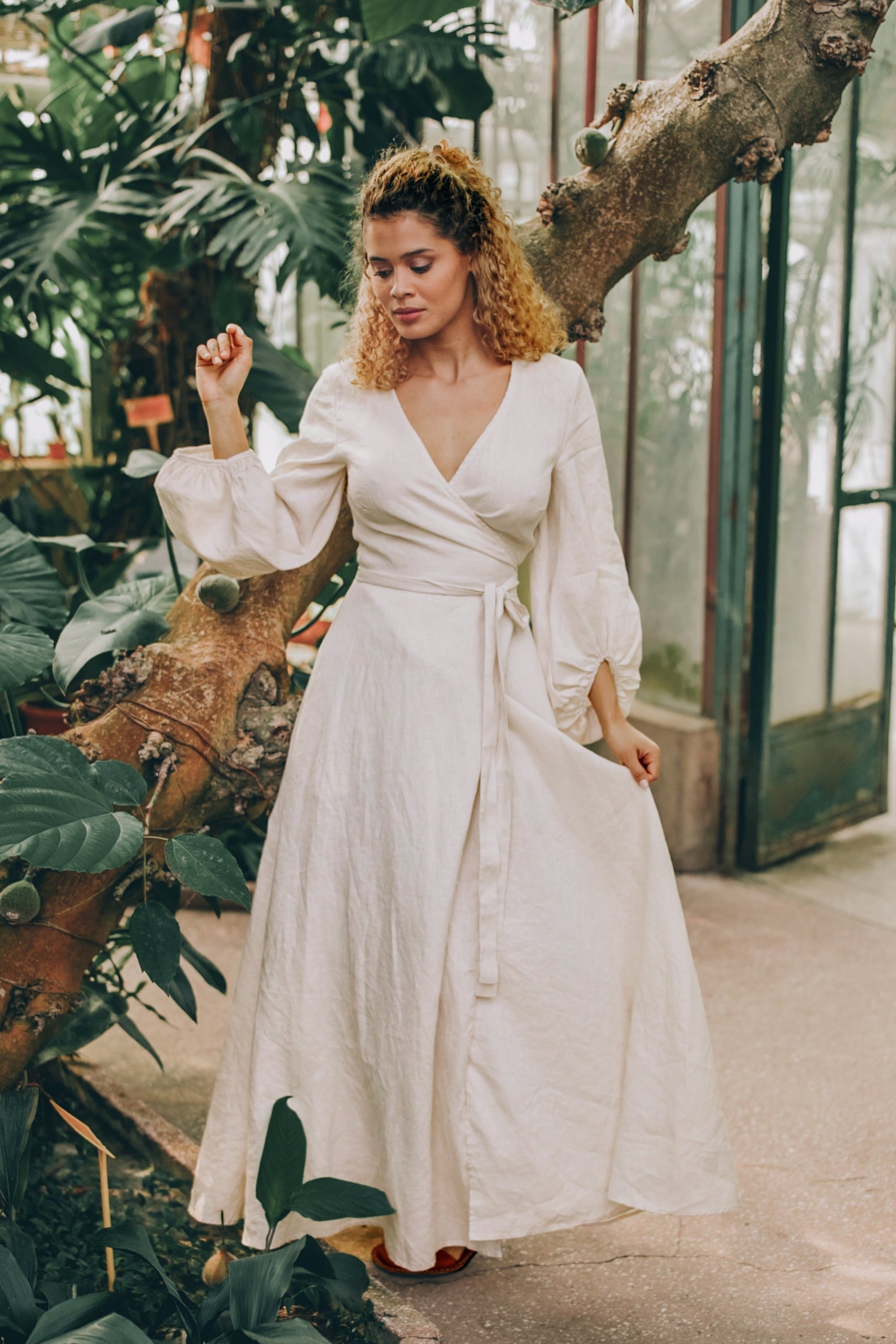 Woman wearing cream linen maxi dress with puff sleeves and wrap tie belt in indoor garden setting