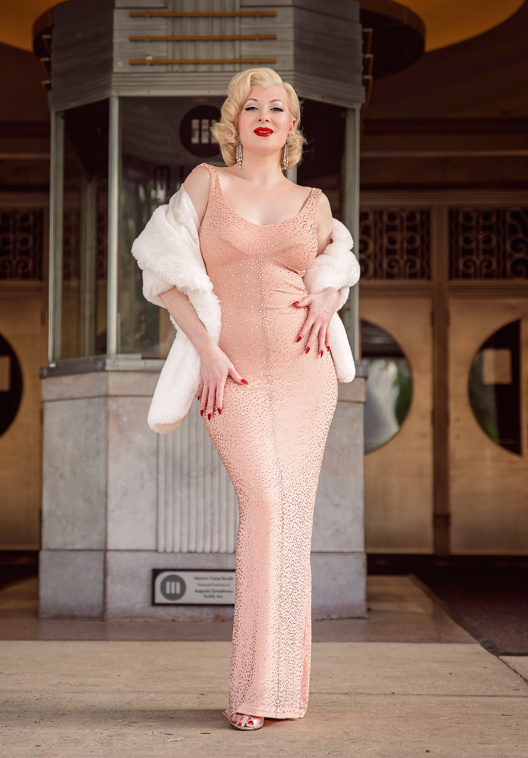 Wearing the HollywoodLand JFK Birthday Dress—a sparkling, form-fitting peach gown inspired by Marilyn Monroe—paired with a white fur stole, vintage waves, and red lipstick, she channels classic Hollywood elegance outside a building.