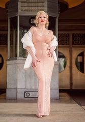 Wearing the HollywoodLand JFK Birthday Dress—a sparkling, form-fitting peach gown inspired by Marilyn Monroe—paired with a white fur stole, vintage waves, and red lipstick, she channels classic Hollywood elegance outside a building.