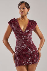 A woman poses confidently in the CRAZY IN LOVE SCALE MINI DRESS, a shiny deep burgundy faux patent leather piece featuring a scale-textured pattern and deep V-neckline, with her hands on her hips against a plain backdrop.
