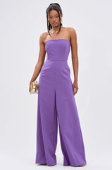 A woman in the BIG BOSS ENERGY WIDE LEG JUMPSUIT IN PURPLE stands confidently with a gold clutch, her straight hair decorated with gold accessories, highlighting the strapless bodice and metallic back zipper against a plain white backdrop.