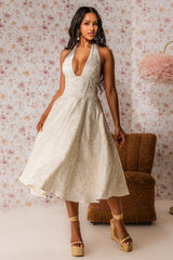 A woman wears the SWEET CAROLINE HALTER MIDI DRESS with gold platform heels, posing before floral wallpaper and a brown vintage chair—an elegant choice for bridal events.