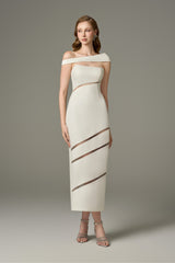 A full-length shot of a woman modeling a serene white off-shoulder, one-sleeve maxi dress with cut-out details and sheer horizontal stripes on the skirt.