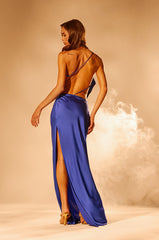 A woman stands in heels, facing away, wearing the RAYNA SATIN MAXI DRESS IN COBALT BLUE. The backless, thigh-high slit design and soft lighting highlight the high-shine satin, creating a dramatic and elegant mood.